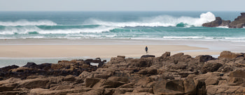 Beach walker strong waves This landscape photograph captures a scene during midday in early spring at Sennen Cove, a well-known coastal location in Cornwall, United Kingdom. The main subject is a solitary beach walker traversing the sand, with strong waves crashing onto the shore and dramatic rocky formations in the foreground. The image highlights the natural beauty of the coast and emphasizes the power of nature, with white surf marking the ocean’s edge. The photograph showcases the rugged landscape characteristic of Sennen Cove and the coastal vistas that define this part of Cornwall.
